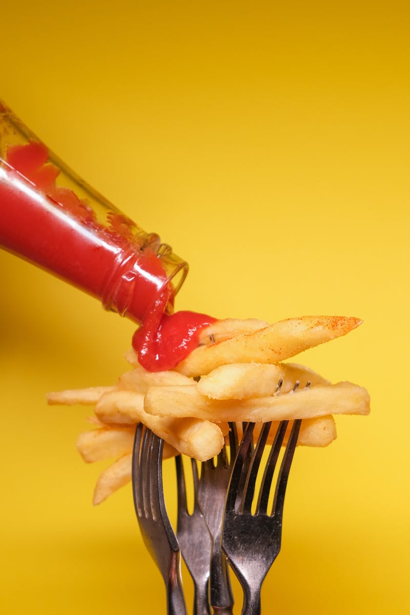 Forks with fried potatoes and bottle with ketchup near yellow background in bright studio